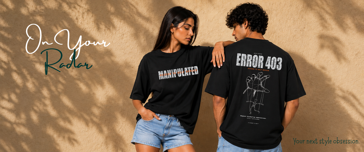 A man and a woman wearing matching black graphic t-shirts pose together against a textured beige wall with leaf shadows. The woman's shirt features the word 'MANIPULATED' in white, while the man's shirt displays 'ERROR 403' with a white line drawing.